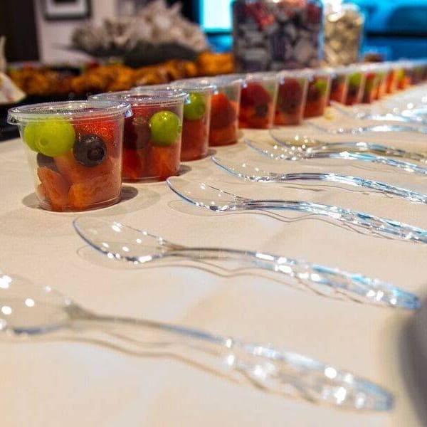 Rows of fruit cups with melon, grapes, and berries arranged alongside clear spoons on a buffet table.