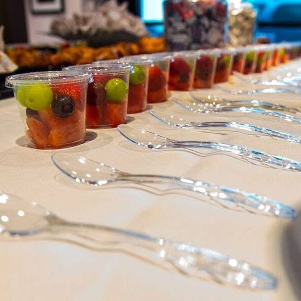 Rows of fruit cups with melon, grapes, and berries arranged alongside clear spoons on a buffet table.