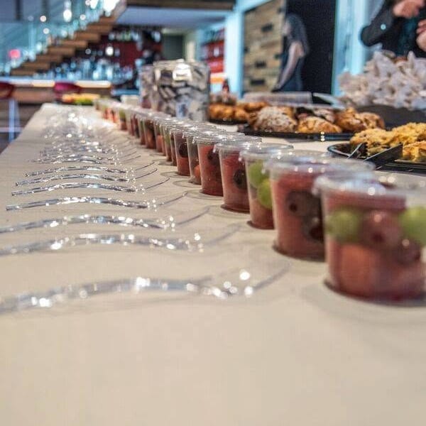 Cups of colorful fruit and dessert samples line a restaurant counter, with pastries, servers, and a modern dining area behind them.