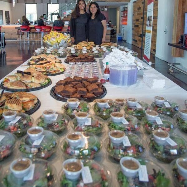 Two women stand behind a table displaying sandwiches, brownies, salads, pastries, and drinks inside a bright café.