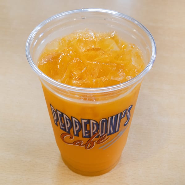A clear plastic cup of iced orange juice with "Pepperoni's Cafe" logo on a light wooden table.
