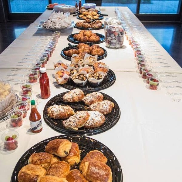 Long buffet table displaying pastries, muffins, fruit cups, sauces, and beverages in a bright event space.