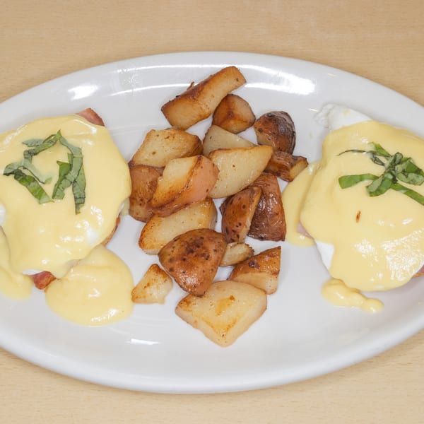 Two eggs Benedict with hollandaise sauce and basil, served with roasted potatoes on a white plate.